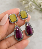 Pair of ruby and yellow sapphire dangle earrings with pavé diamond halos and dark sterling silver settings displayed on hand of a woman